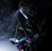 A person in military or tactical gear, wearing a full-face respirator mask and helmet with a mounted light, is operating inside a dark cave or tunnel environment. They are holding a handheld electronic detection device and what appears to be a chemical test kit or sampling material. The scene suggests a CBRN (Chemical, Biological, Radiological, Nuclear) reconnaissance or hazardous material assessment operation.