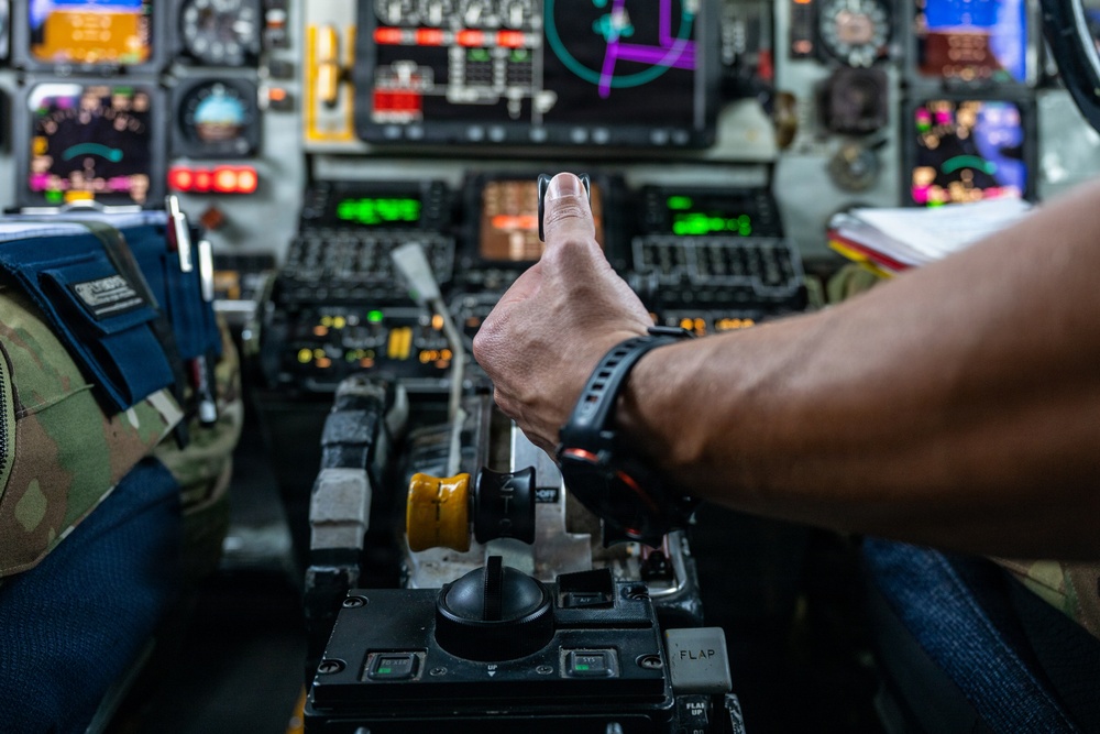 A military aircrew member is giving a thumbs-up gesture while seated in a complex military aircraft cockpit. The cockpit features numerous multifunction displays, analog gauges, and control panels consistent with a large military aircraft. The person is wearing a military flight suit with camouflage pattern and a black sports watch.