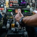 A military aircrew member is giving a thumbs-up gesture while seated in a complex military aircraft cockpit. The cockpit features numerous multifunction displays, analog gauges, and control panels consistent with a large military aircraft. The person is wearing a military flight suit with camouflage pattern and a black sports watch.