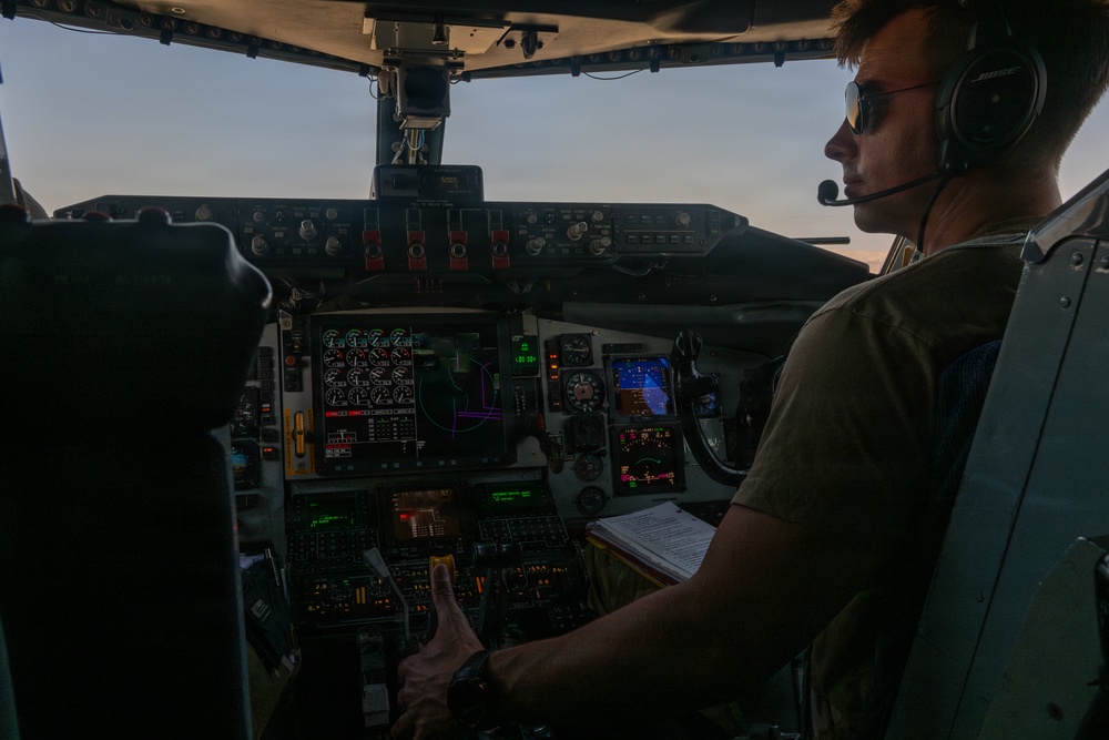 A pilot is seated in the right-side cockpit seat of a large aircraft, operating controls during what appears to be dusk or dawn conditions. The cockpit features a mix of traditional analog instruments and modern digital displays, suggesting a glass cockpit upgrade on an older airframe. The pilot is wearing a Bose aviation headset, sunglasses, and a military-style olive drab t-shirt.