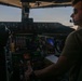 A pilot is seated in the right-side cockpit seat of a large aircraft, operating controls during what appears to be dusk or dawn conditions. The cockpit features a mix of traditional analog instruments and modern digital displays, suggesting a glass cockpit upgrade on an older airframe. The pilot is wearing a Bose aviation headset, sunglasses, and a military-style olive drab t-shirt.