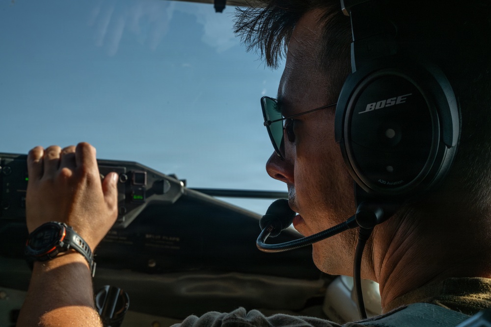 A male pilot or co-pilot is seated in an aircraft cockpit, wearing Bose aviation headphones with a boom microphone and aviator-style sunglasses. He appears to be actively operating or monitoring controls, with his right hand gripping what appears to be a control or instrument panel. The image is taken from a rear-side angle inside the cockpit with a clear blue sky visible through the windscreen.