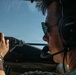 A male pilot or co-pilot is seated in an aircraft cockpit, wearing Bose aviation headphones with a boom microphone and aviator-style sunglasses. He appears to be actively operating or monitoring controls, with his right hand gripping what appears to be a control or instrument panel. The image is taken from a rear-side angle inside the cockpit with a clear blue sky visible through the windscreen.