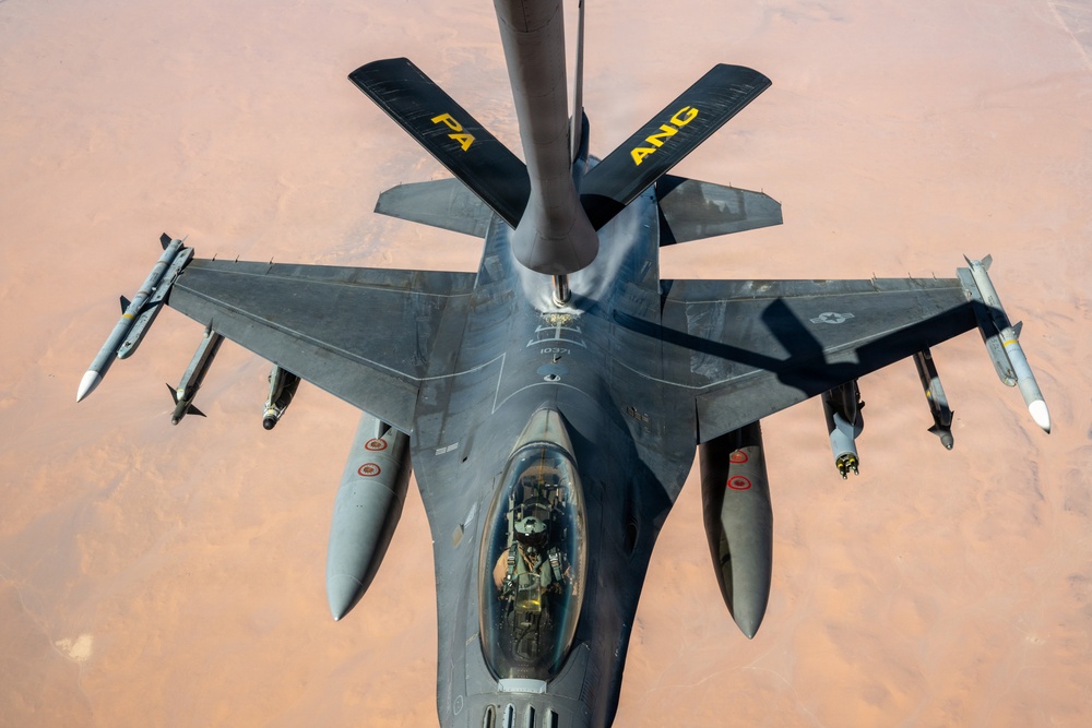An aerial photograph taken from above showing an F-16 Fighting Falcon fighter jet receiving in-flight refueling from a tanker aircraft, visible at the top of the frame. The fighter is fully armed with multiple air-to-air and air-to-ground munitions on its wing pylons. The terrain below appears to be an arid desert landscape, suggesting operations over a Middle Eastern or North African region.