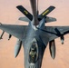An aerial photograph taken from above showing an F-16 Fighting Falcon fighter jet receiving in-flight refueling from a tanker aircraft, visible at the top of the frame. The fighter is fully armed with multiple air-to-air and air-to-ground munitions on its wing pylons. The terrain below appears to be an arid desert landscape, suggesting operations over a Middle Eastern or North African region.