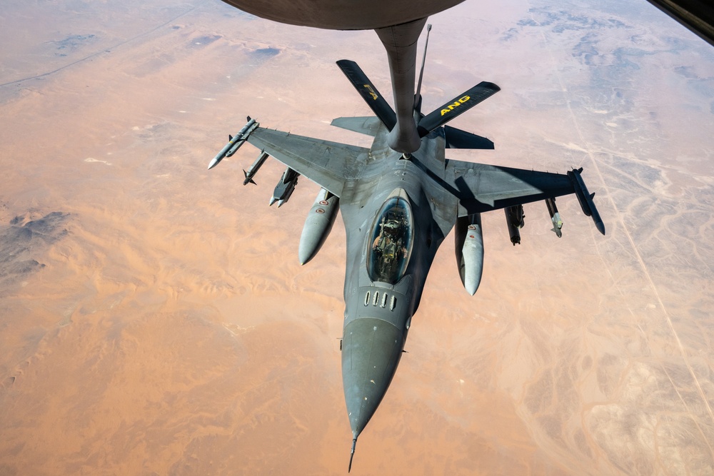 An F-16 Fighting Falcon jet fighter is captured from above during an aerial refueling operation with a tanker aircraft, identifiable by the refueling boom extending downward. The aircraft is flying over an arid desert landscape, likely in the Middle East or a similar region. The tanker bears the marking 'ANG' (Air National Guard) on its tail fins.