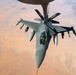An F-16 Fighting Falcon jet fighter is captured from above during an aerial refueling operation with a tanker aircraft, identifiable by the refueling boom extending downward. The aircraft is flying over an arid desert landscape, likely in the Middle East or a similar region. The tanker bears the marking 'ANG' (Air National Guard) on its tail fins.