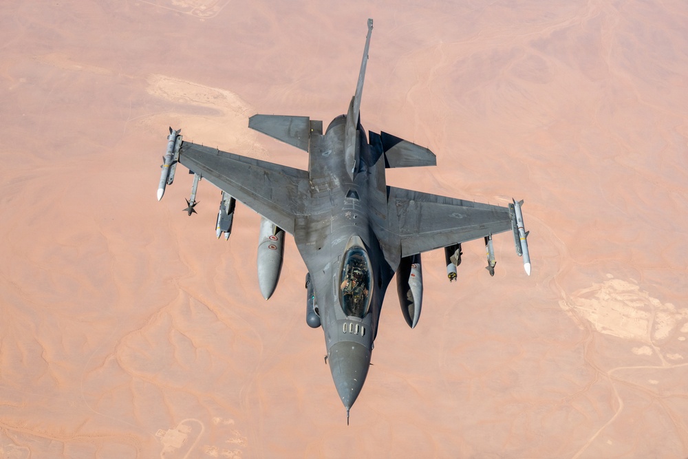 An aerial top-down photograph of a single-seat fighter jet, consistent with an F-16 Fighting Falcon, flying over an arid desert landscape. The aircraft is armed with multiple air-to-air and air-to-ground munitions mounted on its wing pylons. The terrain below appears to be a vast, sparsely populated desert region with reddish-brown earth tones.