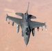 An aerial top-down photograph of a single-seat fighter jet, consistent with an F-16 Fighting Falcon, flying over an arid desert landscape. The aircraft is armed with multiple air-to-air and air-to-ground munitions mounted on its wing pylons. The terrain below appears to be a vast, sparsely populated desert region with reddish-brown earth tones.