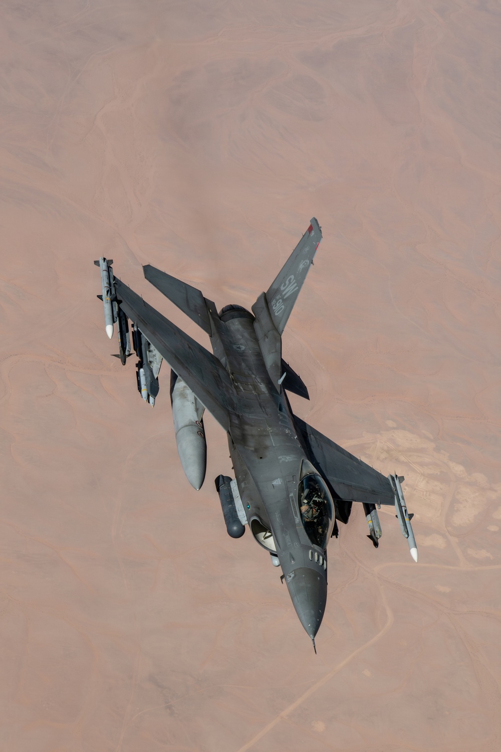 An F-16 Fighting Falcon jet fighter is photographed from above during flight over an arid desert terrain. The aircraft is armed with multiple munitions and carries external fuel tanks, suggesting a combat or patrol mission. The image is taken from another aircraft, likely during a formation flight or aerial refueling operation.