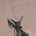An F-16 Fighting Falcon jet fighter is photographed from above during flight over an arid desert terrain. The aircraft is armed with multiple munitions and carries external fuel tanks, suggesting a combat or patrol mission. The image is taken from another aircraft, likely during a formation flight or aerial refueling operation.