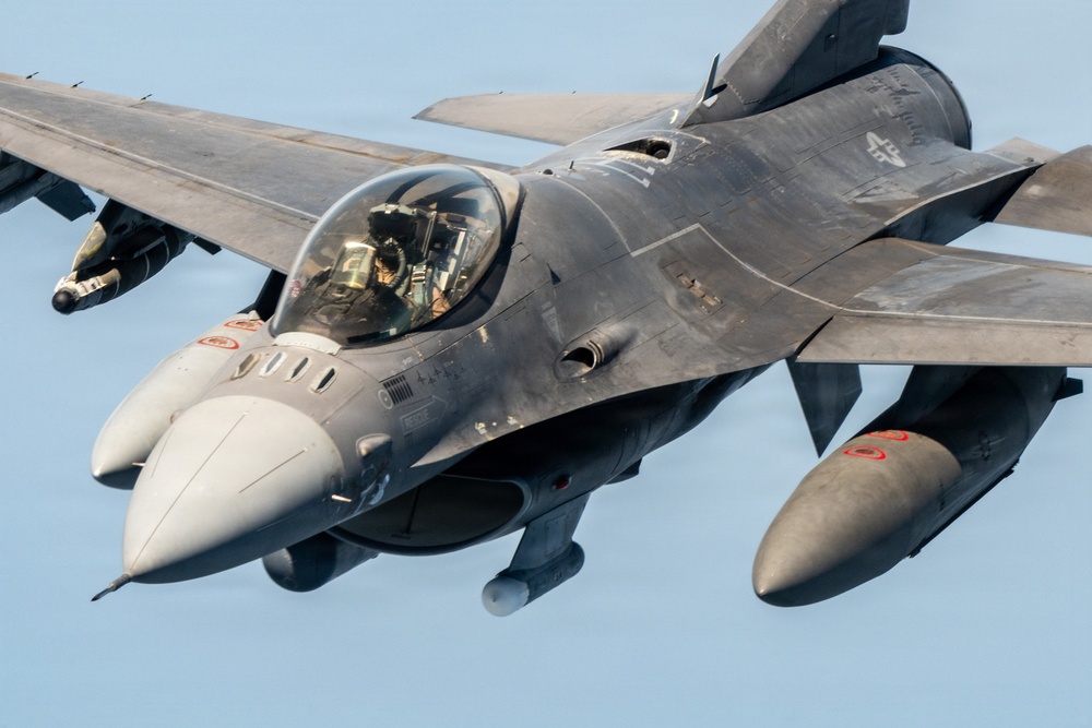 A close-up aerial photograph of an F-16 Fighting Falcon jet fighter in flight against a clear blue sky. The aircraft is shown in a gray camouflage paint scheme with a pilot visible in the cockpit. The image captures fine details of the aircraft's fuselage, canopy, external stores, and markings.