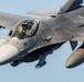 A close-up aerial photograph of an F-16 Fighting Falcon jet fighter in flight against a clear blue sky. The aircraft is shown in a gray camouflage paint scheme with a pilot visible in the cockpit. The image captures fine details of the aircraft's fuselage, canopy, external stores, and markings.