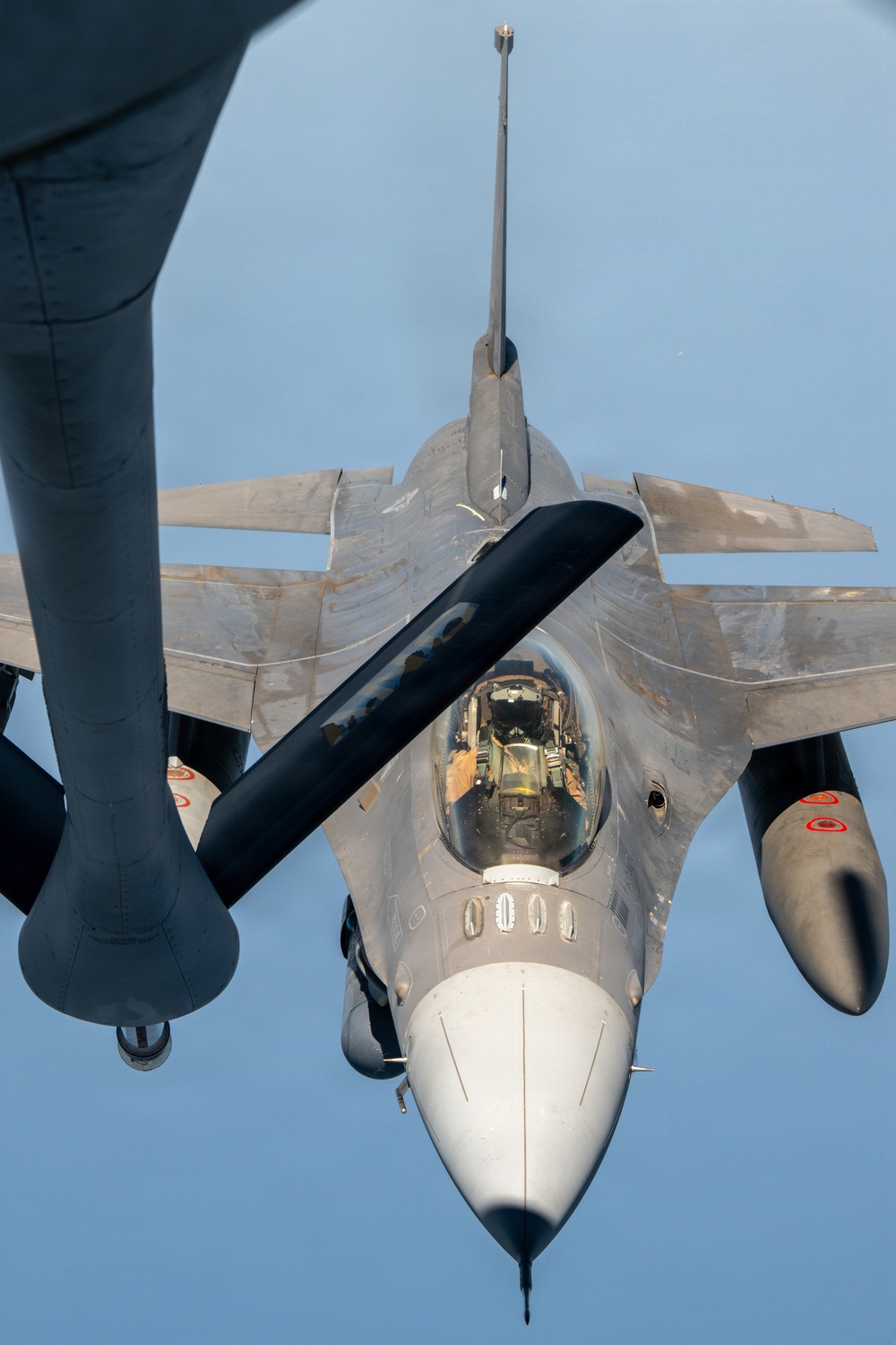 An aerial refueling operation is captured from the perspective of a tanker aircraft looking down at a fighter jet. The receiver aircraft, identifiable as an F-16 Fighting Falcon, is positioned directly below the tanker's refueling boom, which is visible extending diagonally across the frame. The fighter shows signs of heavy use with visible weathering and paint wear on its airframe.