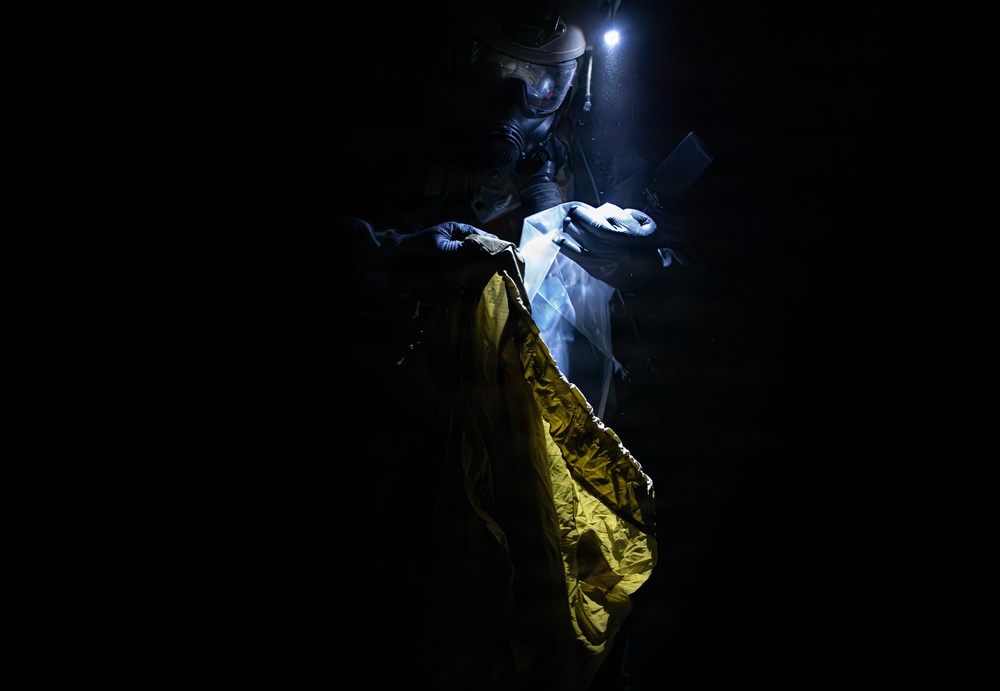 A person wearing protective gear including a helmet with a mounted light, a face shield/visor, and blue gloves is working in near-total darkness. They appear to be handling a yellow garment or material, possibly placing it into or removing it from a clear plastic bag or protective covering. The dramatic low-key lighting creates high contrast between the illuminated subject and the dark background.