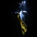 A person wearing protective gear including a helmet with a mounted light, a face shield/visor, and blue gloves is working in near-total darkness. They appear to be handling a yellow garment or material, possibly placing it into or removing it from a clear plastic bag or protective covering. The dramatic low-key lighting creates high contrast between the illuminated subject and the dark background.