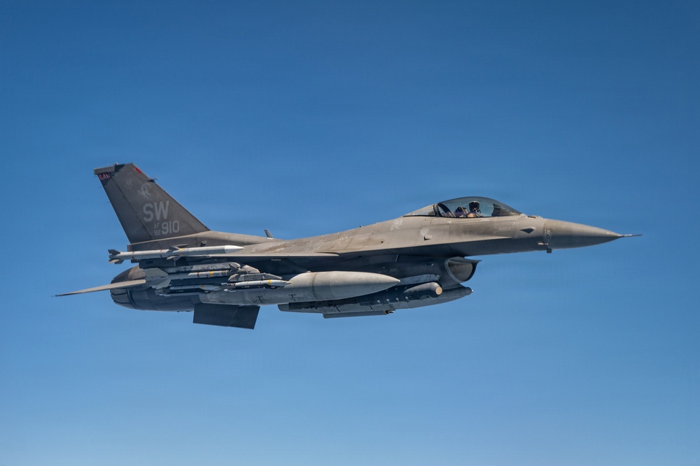 A U.S. Air Force F-16 Fighting Falcon jet fighter is captured in flight against a clear blue sky. The aircraft bears tail markings 'SW 910' indicating it belongs to the 20th Fighter Wing based at Shaw Air Force Base, South Carolina. The aircraft is fully armed with multiple munitions and external fuel/sensor pods mounted under the wings and fuselage.