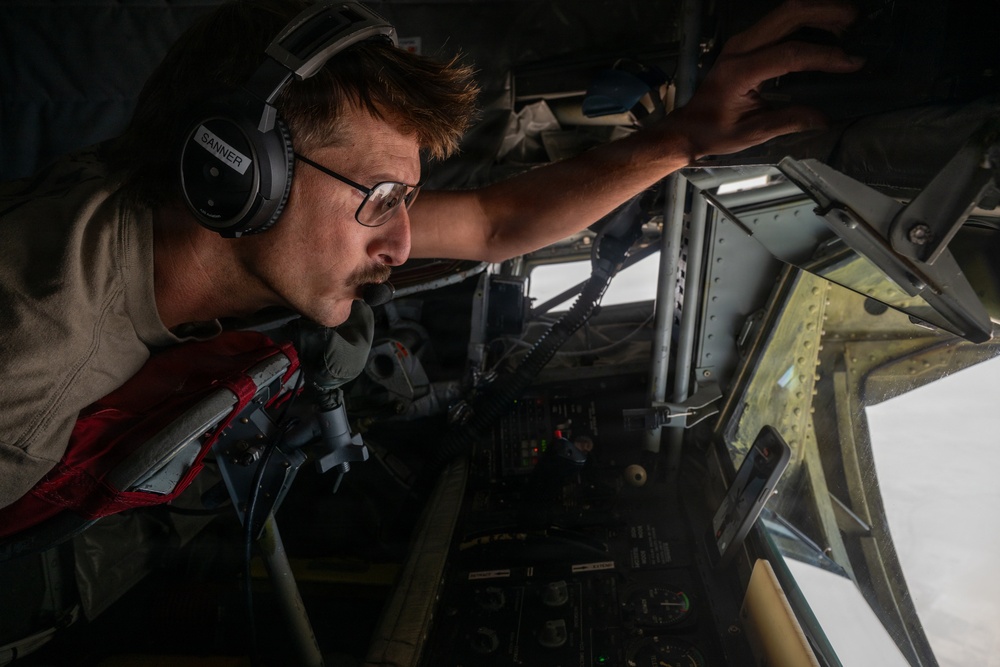 A military aircrew member identified as 'SANNER' on his headset is leaning forward intently inside an aircraft cockpit, appearing to look out through a forward window or inspect equipment. He is wearing aviation headphones, glasses, a tan flight suit, and a red survival vest. The image is taken from a low angle inside the aircraft, capturing both the operator and the complex cockpit instrumentation.