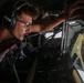 A military aircrew member identified as 'SANNER' on his headset is leaning forward intently inside an aircraft cockpit, appearing to look out through a forward window or inspect equipment. He is wearing aviation headphones, glasses, a tan flight suit, and a red survival vest. The image is taken from a low angle inside the aircraft, capturing both the operator and the complex cockpit instrumentation.