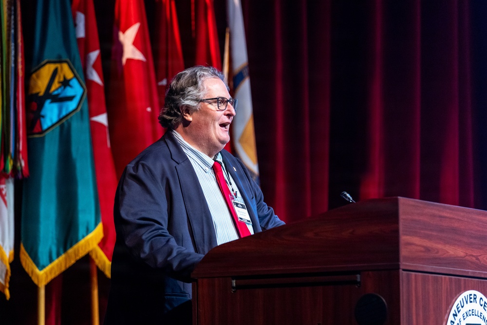 A civilian male speaker addresses an audience from a wooden podium bearing partial text reading 'NEUVER CE[NTER] OF EXCELLEN[CE]', consistent with the Maneuver Center of Excellence. He is dressed in a navy blazer, striped shirt, and red tie, and wears a lanyard with a badge. Multiple military unit flags and a red curtain backdrop are visible behind him.