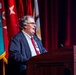 A civilian male speaker addresses an audience from a wooden podium bearing partial text reading 'NEUVER CE[NTER] OF EXCELLEN[CE]', consistent with the Maneuver Center of Excellence. He is dressed in a navy blazer, striped shirt, and red tie, and wears a lanyard with a badge. Multiple military unit flags and a red curtain backdrop are visible behind him.