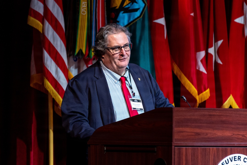 A man in a navy blue blazer and red tie is speaking at a wooden podium during a formal event. The podium bears partial text reading 'EUVER C' suggesting a military installation or conference venue. Multiple military and organizational flags are displayed prominently in the background.