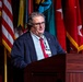 A man in a navy blue blazer and red tie is speaking at a wooden podium during a formal event. The podium bears partial text reading 'EUVER C' suggesting a military installation or conference venue. Multiple military and organizational flags are displayed prominently in the background.