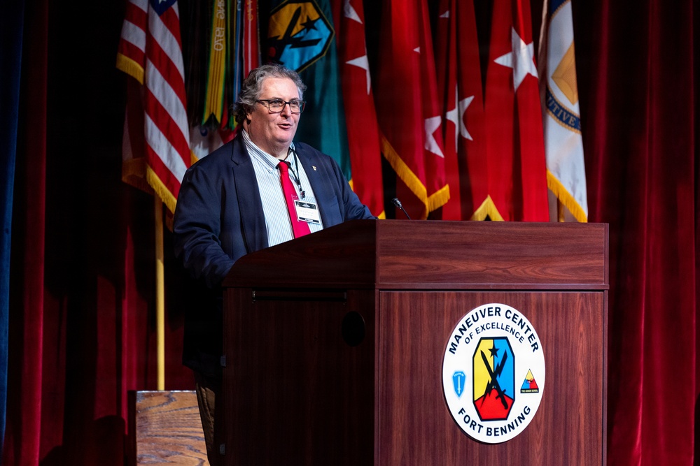 A civilian male speaker addresses an audience from a wooden podium bearing the official seal of the Maneuver Center of Excellence at Fort Benning. He is wearing a dark blazer, red tie, and glasses, with a lanyard/badge visible around his neck. Multiple military and U.S. flags are displayed prominently on stage behind him.