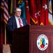 A civilian male speaker addresses an audience from a wooden podium bearing the official seal of the Maneuver Center of Excellence at Fort Benning. He is wearing a dark blazer, red tie, and glasses, with a lanyard/badge visible around his neck. Multiple military and U.S. flags are displayed prominently on stage behind him.