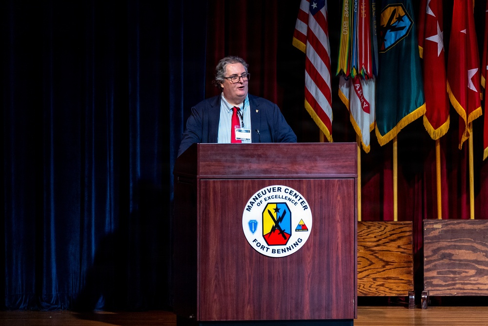A civilian male speaker addresses an audience from a wooden podium at Fort Benning's Maneuver Center of Excellence. He is wearing a dark blazer, striped shirt, red tie, and glasses, with a lanyard/badge visible. Multiple military unit flags and the U.S. flag are displayed on stage behind him.