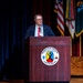 A civilian male speaker addresses an audience from a wooden podium at Fort Benning's Maneuver Center of Excellence. He is wearing a dark blazer, striped shirt, red tie, and glasses, with a lanyard/badge visible. Multiple military unit flags and the U.S. flag are displayed on stage behind him.