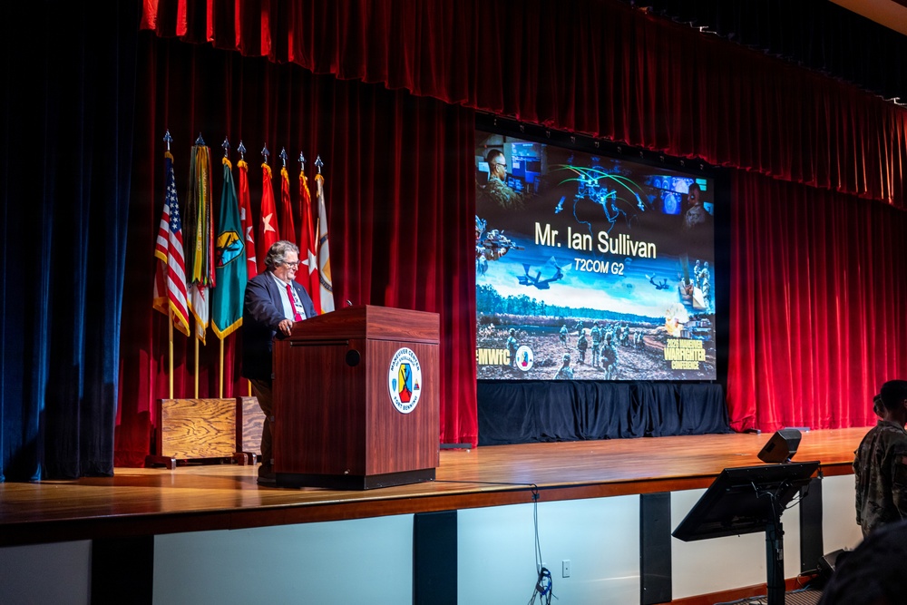 A man identified as Mr. Ian Sullivan, representing T2COM G2, is delivering a presentation at a military conference podium on a stage. The event appears to be the 'Force Maneuver Warfighter Conference' (MWIG), indicated by branding visible on the projection screen. Multiple military flags are displayed behind the speaker on the stage.