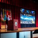 A man identified as Mr. Ian Sullivan, representing T2COM G2, is delivering a presentation at a military conference podium on a stage. The event appears to be the 'Force Maneuver Warfighter Conference' (MWIG), indicated by branding visible on the projection screen. Multiple military flags are displayed behind the speaker on the stage.