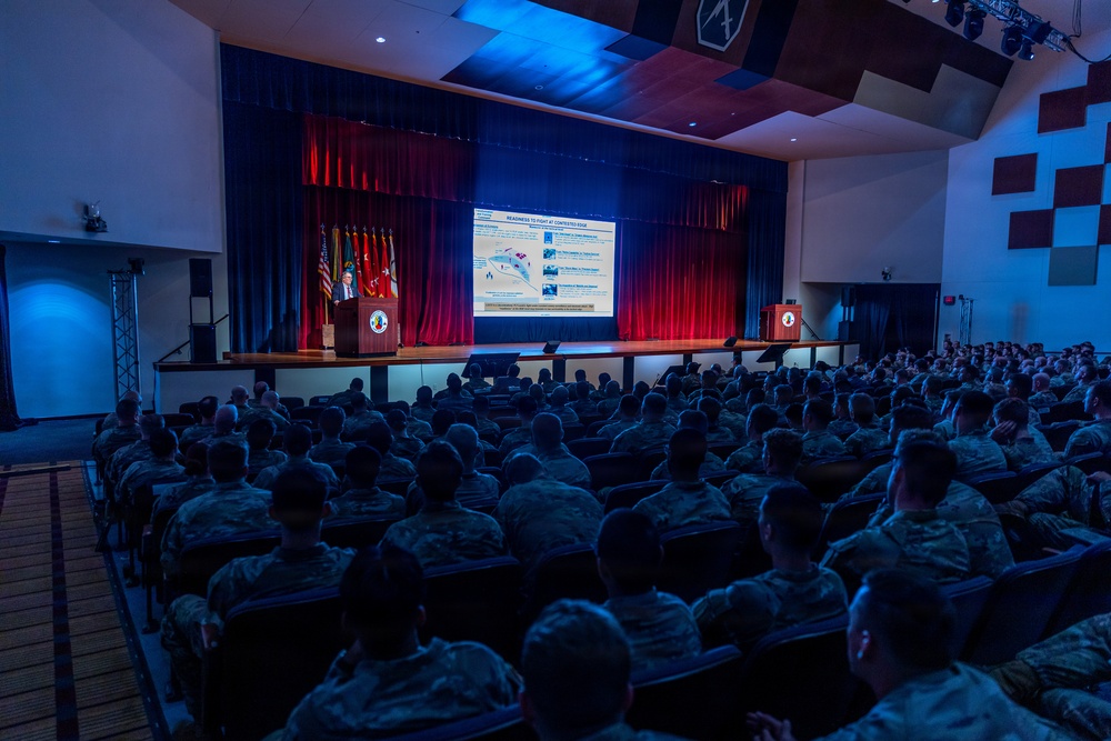 A civilian speaker addresses a large audience of military personnel in camouflage uniforms inside a formal auditorium or theater. The speaker stands at a podium on a stage flanked by American and military unit flags, with a large projection screen displaying a briefing slide titled 'READINESS TO FIGHT AT CONTESTED EDGE.' The venue appears to be a military installation auditorium with professional stage lighting and acoustic wall panels.