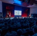A civilian speaker addresses a large audience of military personnel in camouflage uniforms inside a formal auditorium or theater. The speaker stands at a podium on a stage flanked by American and military unit flags, with a large projection screen displaying a briefing slide titled 'READINESS TO FIGHT AT CONTESTED EDGE.' The venue appears to be a military installation auditorium with professional stage lighting and acoustic wall panels.