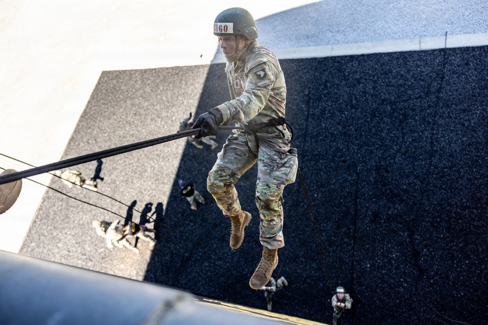 A soldier in full military camouflage uniform is rappelling down a tall dark wall structure, gripping a rope while suspended in mid-air. The soldier wears a helmet with visible labeling, tactical gloves, and tan combat boots. Additional personnel are visible at the base of the structure, suggesting a supervised military training exercise.
