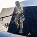 A soldier in full military camouflage uniform is rappelling down a tall dark wall structure, gripping a rope while suspended in mid-air. The soldier wears a helmet with visible labeling, tactical gloves, and tan combat boots. Additional personnel are visible at the base of the structure, suggesting a supervised military training exercise.