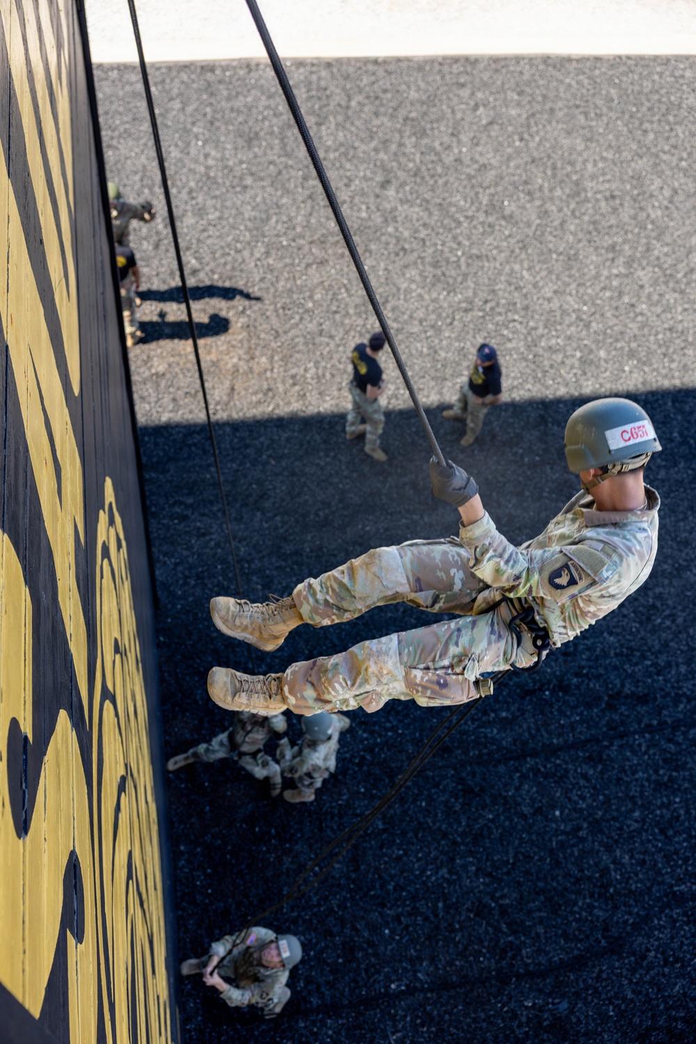 A military or ROTC cadet in MultiCam/OCP camouflage uniform is rappelling down a tall training tower wall painted black and yellow. The image is taken from an elevated angle looking down, showing the rappeller suspended on ropes with feet planted against the wall. Several other uniformed personnel are visible on the ground below, observing or waiting their turn.