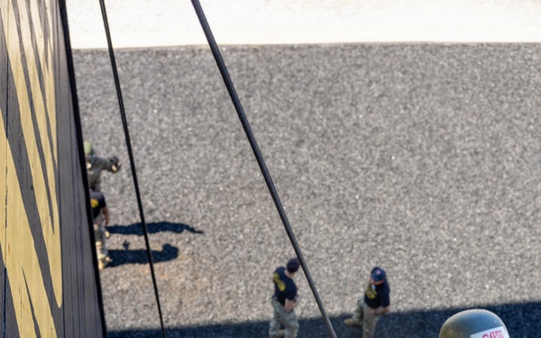 A military or ROTC cadet in MultiCam/OCP camouflage uniform is rappelling down a tall training tower wall painted black and yellow. The image is taken from an elevated angle looking down, showing the rappeller suspended on ropes with feet planted against the wall. Several other uniformed personnel are visible on the ground below, observing or waiting their turn.