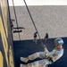 A military or ROTC cadet in MultiCam/OCP camouflage uniform is rappelling down a tall training tower wall painted black and yellow. The image is taken from an elevated angle looking down, showing the rappeller suspended on ropes with feet planted against the wall. Several other uniformed personnel are visible on the ground below, observing or waiting their turn.