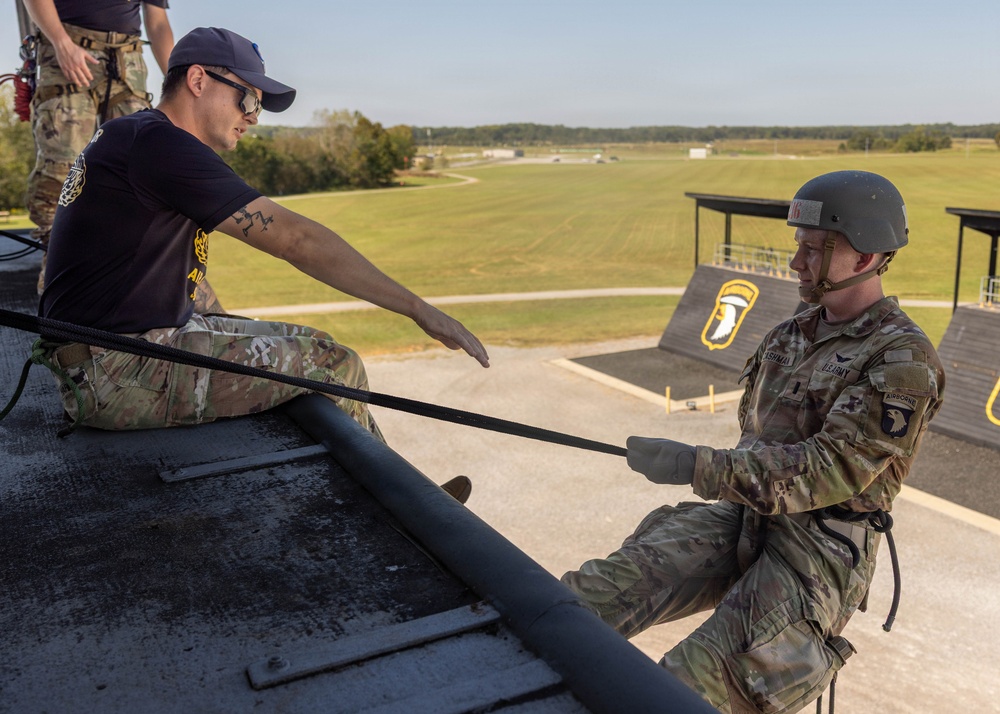 A U.S. Army Airborne soldier wearing a helmet and OCP camouflage uniform is rappelling or preparing to descend from the edge of an elevated training tower. An instructor in a navy blue t-shirt and OCP pants is seated at the edge guiding the soldier, while a third person in full gear stands in the background. The 101st Airborne Division screaming eagle insignia is visible on a structure in the background, indicating this is likely Fort Campbell or a related training facility.