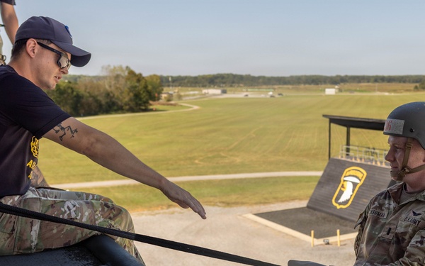 A U.S. Army Airborne soldier wearing a helmet and OCP camouflage uniform is rappelling or preparing to descend from the edge of an elevated training tower. An instructor in a navy blue t-shirt and OCP pants is seated at the edge guiding the soldier, while a third person in full gear stands in the background. The 101st Airborne Division screaming eagle insignia is visible on a structure in the background, indicating this is likely Fort Campbell or a related training facility.