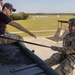 A U.S. Army Airborne soldier wearing a helmet and OCP camouflage uniform is rappelling or preparing to descend from the edge of an elevated training tower. An instructor in a navy blue t-shirt and OCP pants is seated at the edge guiding the soldier, while a third person in full gear stands in the background. The 101st Airborne Division screaming eagle insignia is visible on a structure in the background, indicating this is likely Fort Campbell or a related training facility.