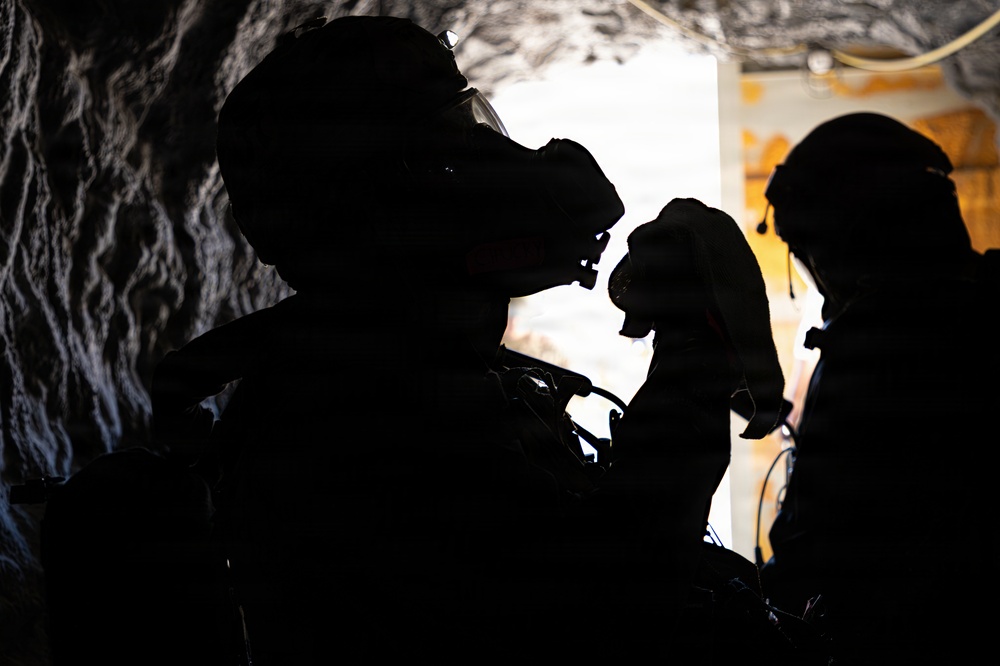 The image shows two heavily silhouetted figures in what appears to be a confined or underground environment, both wearing tactical helmets and specialized equipment including what appears to be night vision or gas mask apparatus. The strong backlight creates dramatic silhouettes, obscuring most detail. The setting suggests a military, law enforcement, or emergency response context.