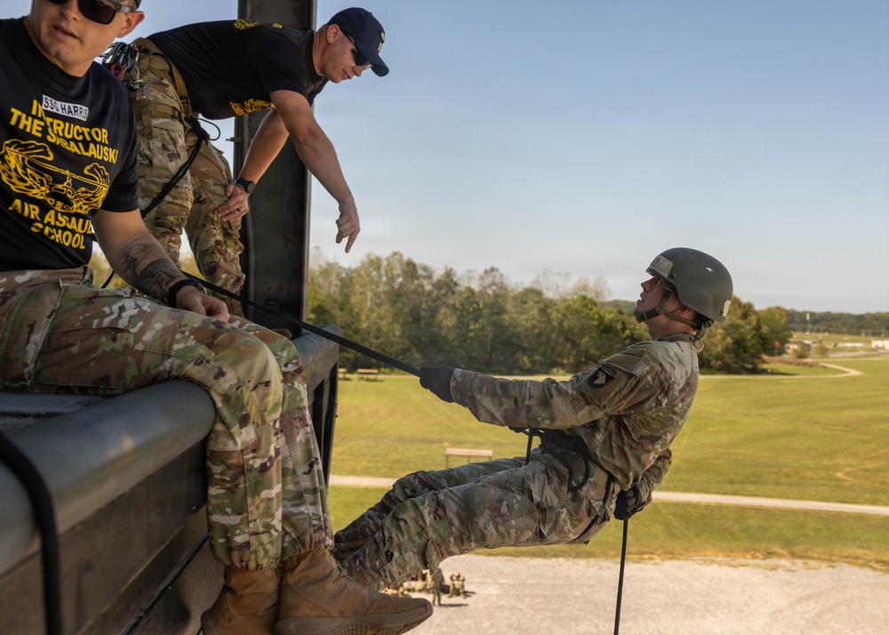 Military personnel are conducting rappelling training at what appears to be the Air Assault School, likely at Fort Campbell, Kentucky. An instructor wearing a black 'Air Assault School' t-shirt and another in camouflage uniform are supervising a student who is leaning back over the edge of an elevated platform secured by a rappel rope. The training appears to be a controlled tower rappel exercise in clear daylight conditions.