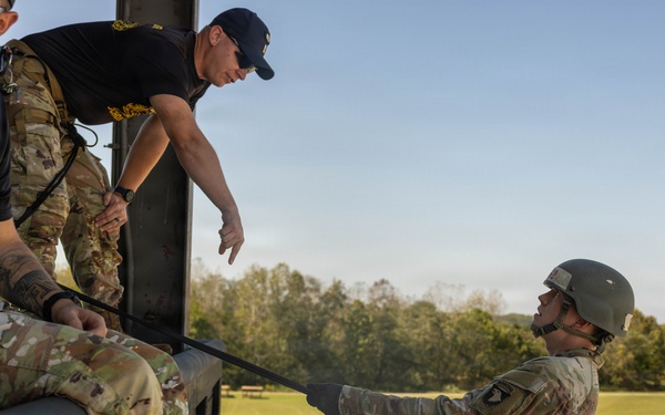 Military personnel are conducting rappelling training at what appears to be the Air Assault School, likely at Fort Campbell, Kentucky. An instructor wearing a black 'Air Assault School' t-shirt and another in camouflage uniform are supervising a student who is leaning back over the edge of an elevated platform secured by a rappel rope. The training appears to be a controlled tower rappel exercise in clear daylight conditions.