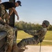 Military personnel are conducting rappelling training at what appears to be the Air Assault School, likely at Fort Campbell, Kentucky. An instructor wearing a black 'Air Assault School' t-shirt and another in camouflage uniform are supervising a student who is leaning back over the edge of an elevated platform secured by a rappel rope. The training appears to be a controlled tower rappel exercise in clear daylight conditions.