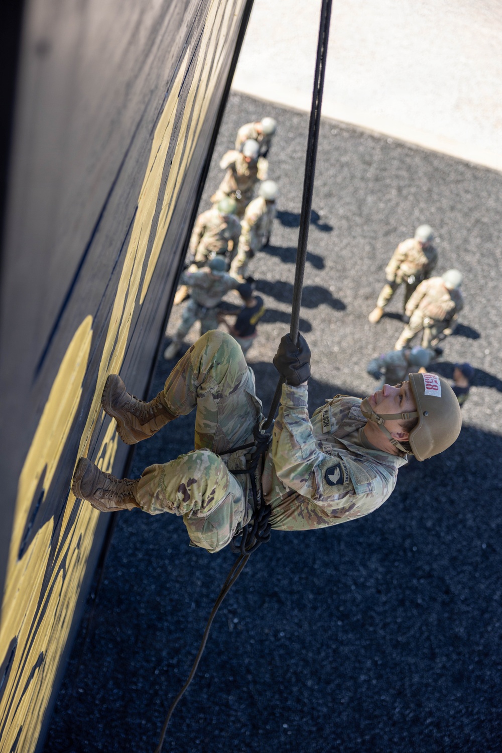 A military service member in OCP (Operational Camouflage Pattern) uniform is rappelling down a tall dark-colored wall during what appears to be a training exercise. The photo is taken from above, providing a dramatic top-down perspective showing the rappeller mid-descent with several other uniformed personnel visible on the ground below. The individual wears a helmet with a numbered identification tag (058) and a name tape reading 'WILSON'.
