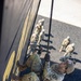 A military service member in OCP (Operational Camouflage Pattern) uniform is rappelling down a tall dark-colored wall during what appears to be a training exercise. The photo is taken from above, providing a dramatic top-down perspective showing the rappeller mid-descent with several other uniformed personnel visible on the ground below. The individual wears a helmet with a numbered identification tag (058) and a name tape reading 'WILSON'.