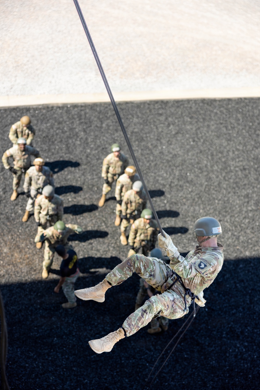 A military soldier in full combat uniform and helmet is rappelling down a rope from an elevated position, viewed from above. A group of approximately 10-12 soldiers in camouflage uniforms and helmets are gathered on the ground below, observing the descent. This appears to be a military rappelling or airborne training exercise.
